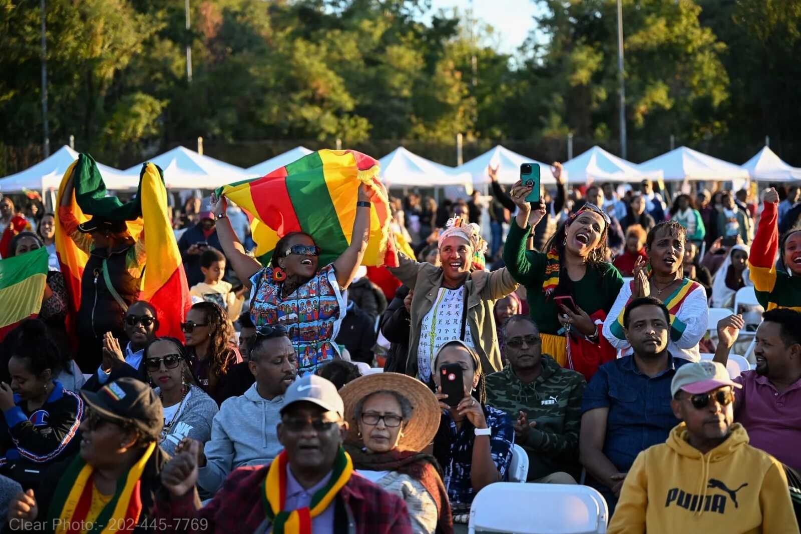 Crowd at an outdoor festival waving colorful flags and cheering, with white tents in the background.