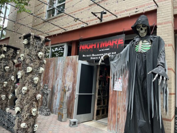 Entrance to a Halloween pop-up bar with a tall skeletal figure in a black cloak guarding the doorway, Nightmare on the Avenue banner above.