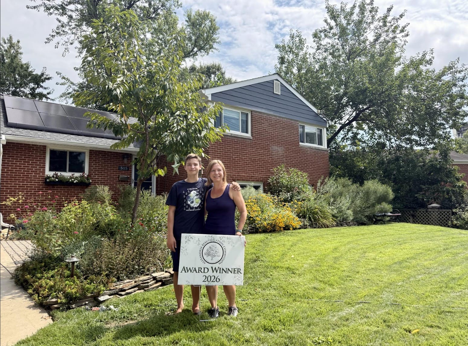 A woman and teenage boy stand in a front yard holding an 'Award Winner 2026' sign, with a brick house and garden behind them.