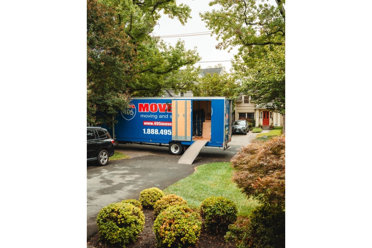 Blue moving truck parked in a suburban driveway with a ramp extended, boxes visible inside as it’s being loaded. Green trees and houses line the street.
