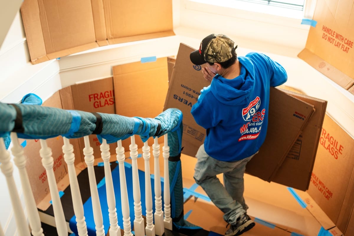 Mover wearing a blue hoodie carries a large cardboard box up a stairwell surrounded by fragile-glass boxes labeled for packing.