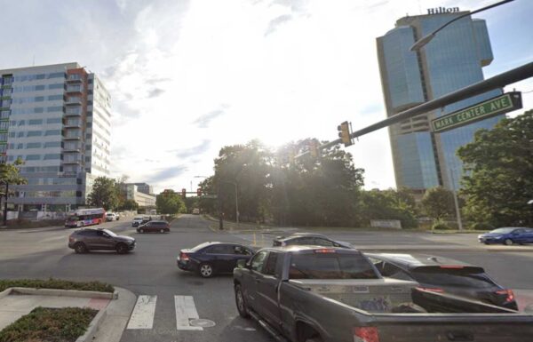 Busy urban intersection with several cars waiting at a cross street, modern glass buildings in the background including a tall Hilton hotel on the right.