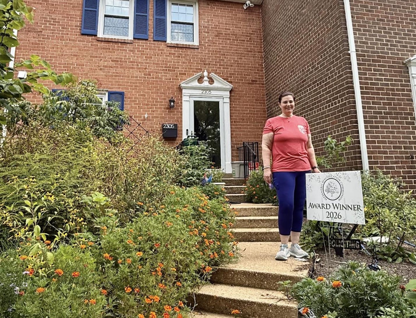 Woman in pink shirt and purple leggings standing on the front steps of a brick house with a sign reading 'Award Winner 2026' in a garden bed of orange flowers in the foreground.