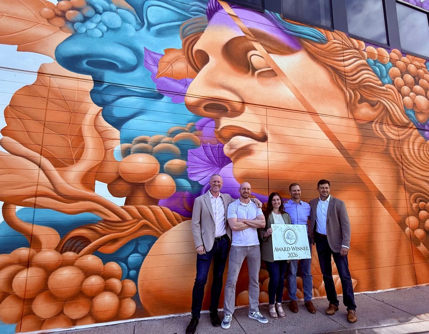 Five adults stand together in front of a vibrant mural featuring a large stylized woman's face with orange-blue abstract shapes, holding a sign that reads 'Award Winner 2026'.