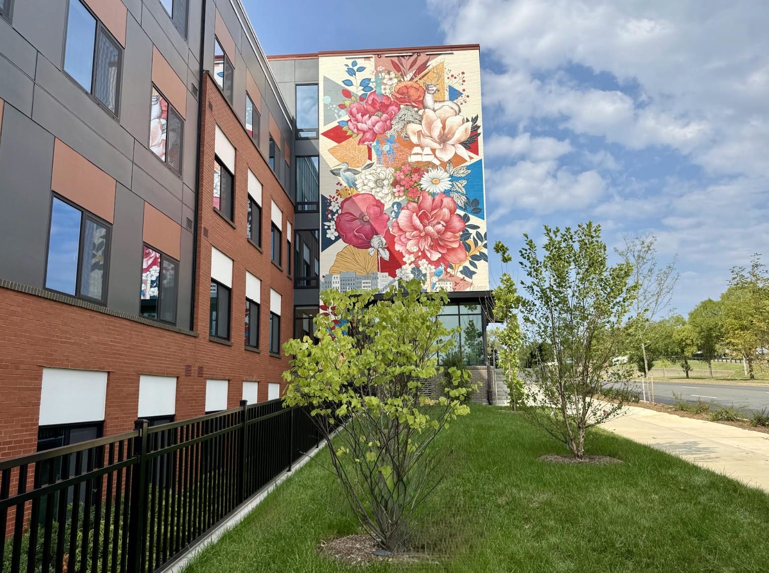 Modern brick and gray-panel building with a large colorful flower mural on its side, green lawn and trees in foreground.