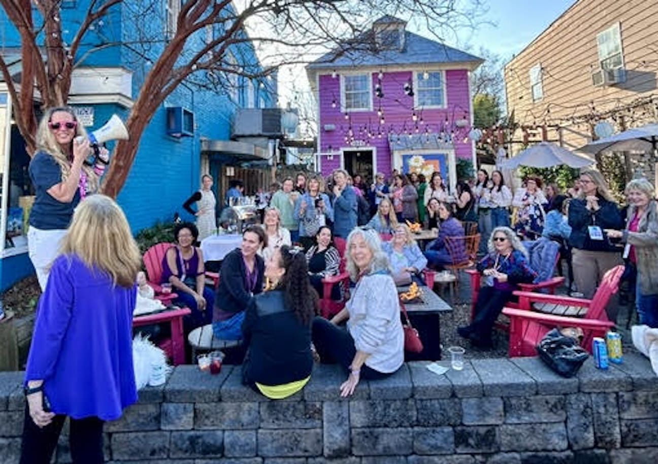 Speaker with a megaphone addressing a large outdoor crowd in a colorful courtyard with pink and blue buildings and string lights.