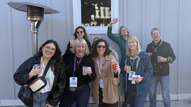 A group of eight adults smiling and posing together outside a light-blue wall, some waving, holding drinks and wearing lanyards with badges.